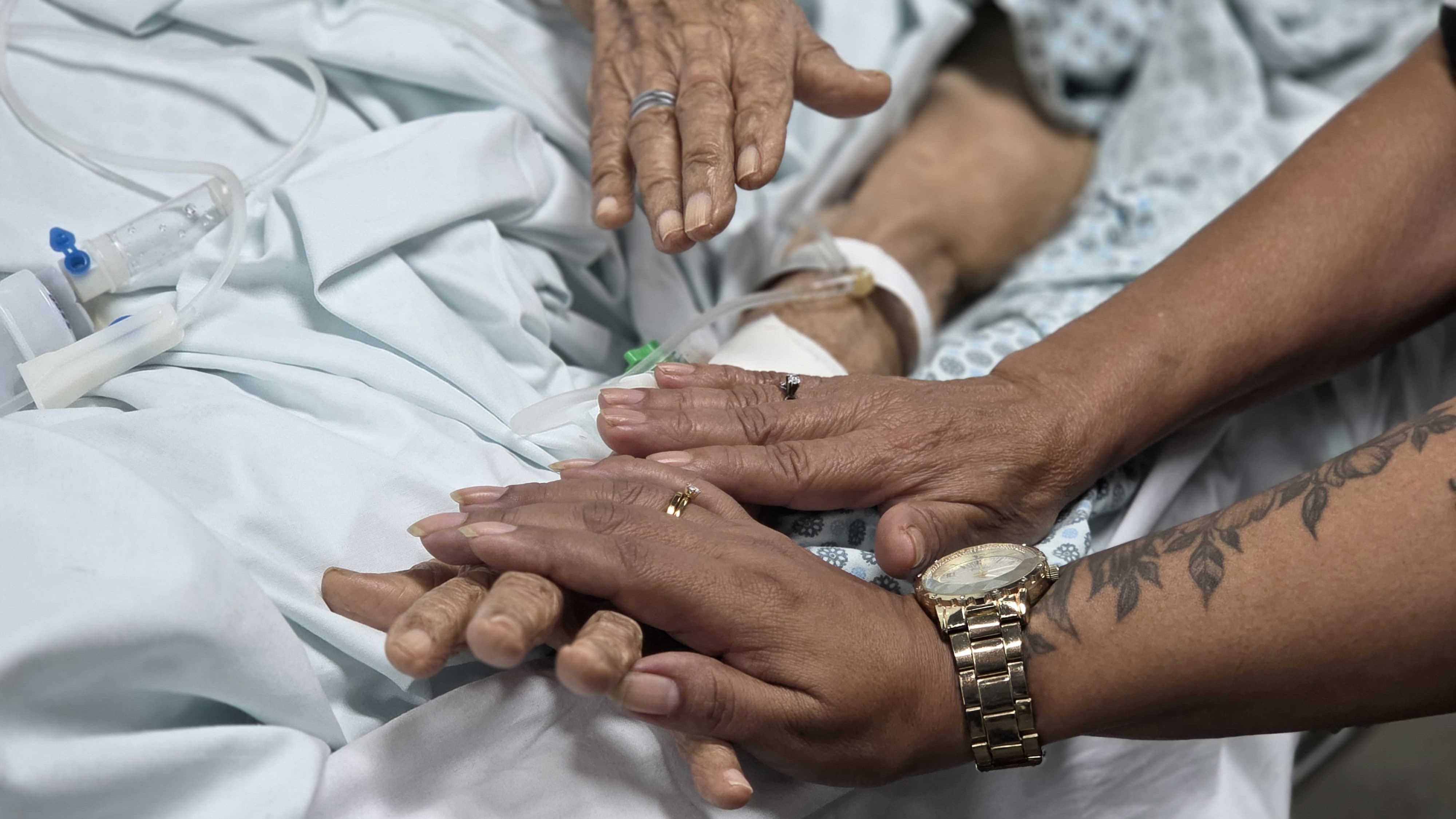 a foto mostra mãos entrelaçadas de um idoso e uma pessoal mais jovem numa cama de hospital